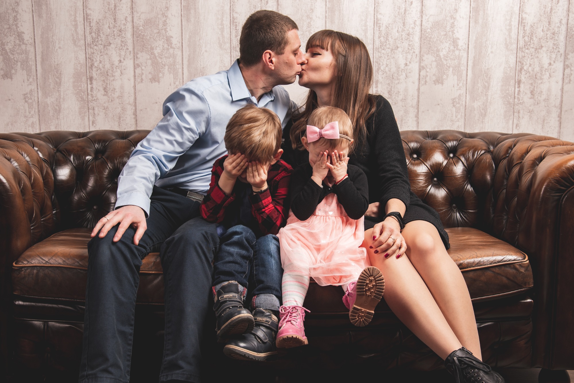 Rodzinne ujęcie na skórzanej sofie: rodzice całują się, podczas gdy dzieci zakrywają twarze dłońmi, ukazując swoje buty. Dziewczynka ma różową sukienkę i kokardę.