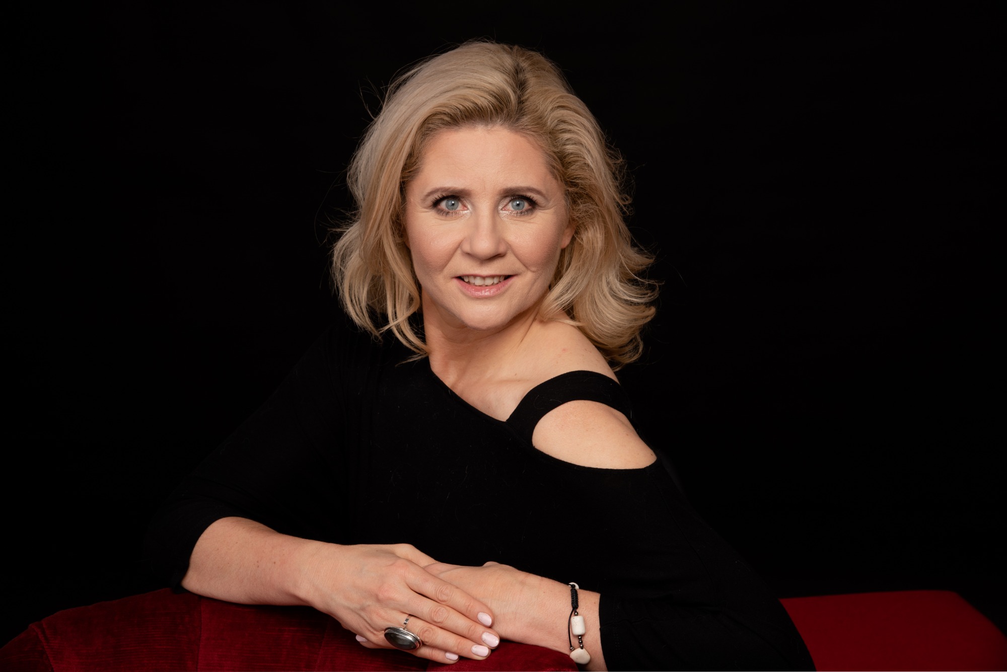 Portret uśmiechniętej blondynki w czarnej bluzce z wycięciem na ramieniu, opierającej ręce na czerwonym aksamicie na czarnym tle.