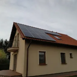 Dom jednorodzinny z zamontowanymi panelami fotowoltaicznymi na dachu pokrytym czerwoną dachówką, widok z dołu na pochmurne niebo.