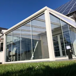 Szklana zabudowa tarasu z beżową ramą, przylegająca do domu z panelami słonecznymi na dachu, widok z zewnątrz na trawnik.