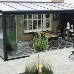 Zadaszony taras z przesuwnymi szklanymi ścianami, wyposażony w stół z krzesłami, regał z dekoracjami i ozdobną posadzkę w geometryczny wzór.