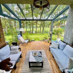 FHU B&B Bartosz Ginko-Beszterda - Zadaszony taras z drewnianą podłogą i szklanymi ścianami, wyposażony w sofy z jasnymi poduszkami, fotel, stolik kawowy z dekoracjami i dywan. Widok na zielony ogród przez szklane ściany. Na jednej...