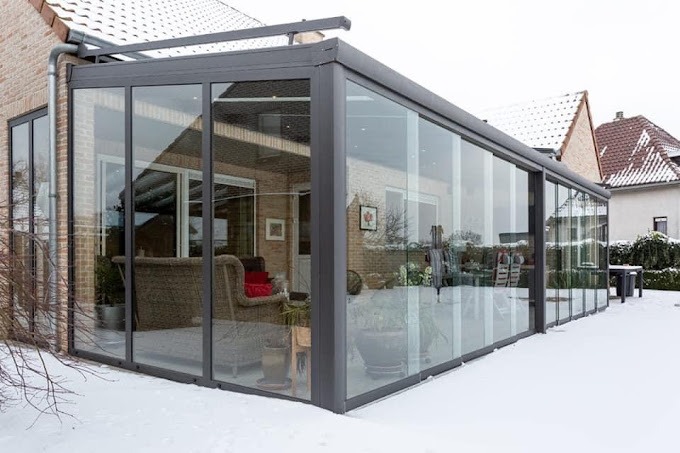 Szklana zabudowa tarasu z metalową konstrukcją w zimowej scenerii, widoczne meble ogrodowe i dekoracje.
