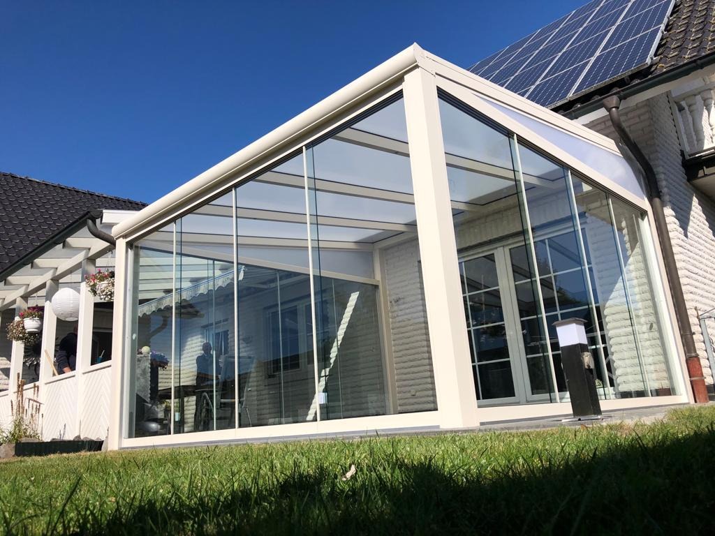 Szklana zabudowa tarasu z beżową ramą, przylegająca do domu z panelami słonecznymi na dachu, widok z zewnątrz na trawnik.