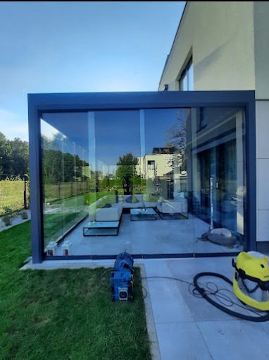 Nowoczesna przeszklona pergola z szarą ramą przylegająca do domu, z widocznymi meblami wypoczynkowymi wewnątrz, oraz sprzętem czyszczącym na zewnątrz.
