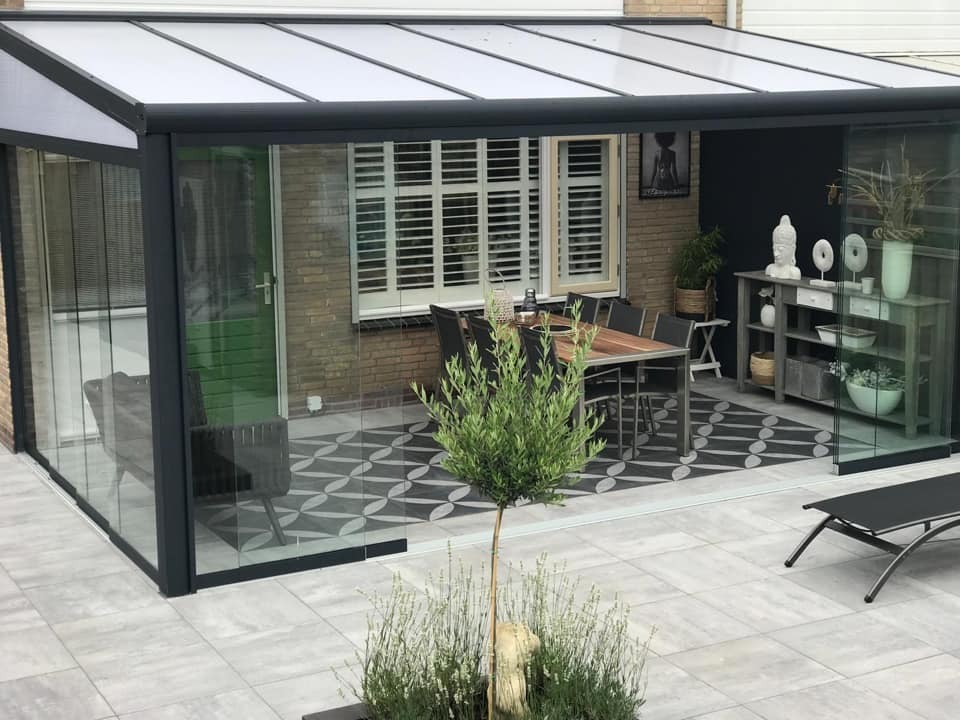 Zadaszony taras z przesuwnymi szklanymi ścianami, wyposażony w stół z krzesłami, regał z dekoracjami i ozdobną posadzkę w geometryczny wzór.