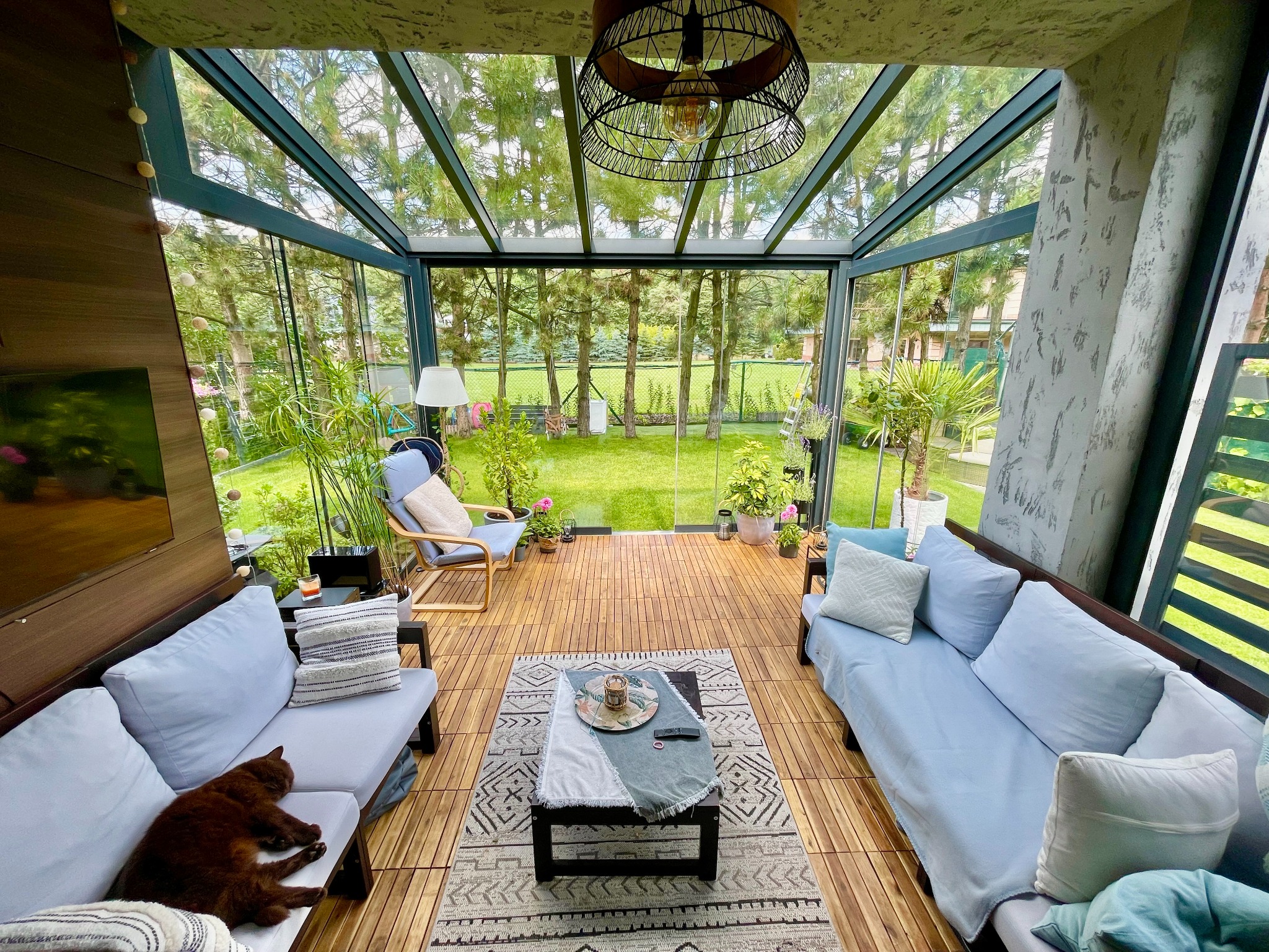 Zadaszony taras z drewnianą podłogą i szklanymi ścianami, wyposażony w sofy z jasnymi poduszkami, fotel, stolik kawowy z dekoracjami i dywan. Widok na zielony ogród przez szklane ściany. Na jednej...