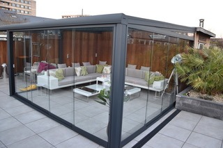 Szklana zabudowa tarasu z szarą ramą, widoczne meble wypoczynkowe wewnątrz, rośliny doniczkowe i fragment otoczenia miejskiego.