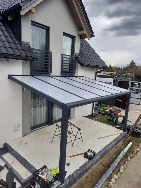 Nowoczesna pergola metalowa z przezroczystym zadaszeniem poliwęglanowym, zamontowana przy domu z balkonami, widoczne narzędzia i materiały budowlane na tarasie.