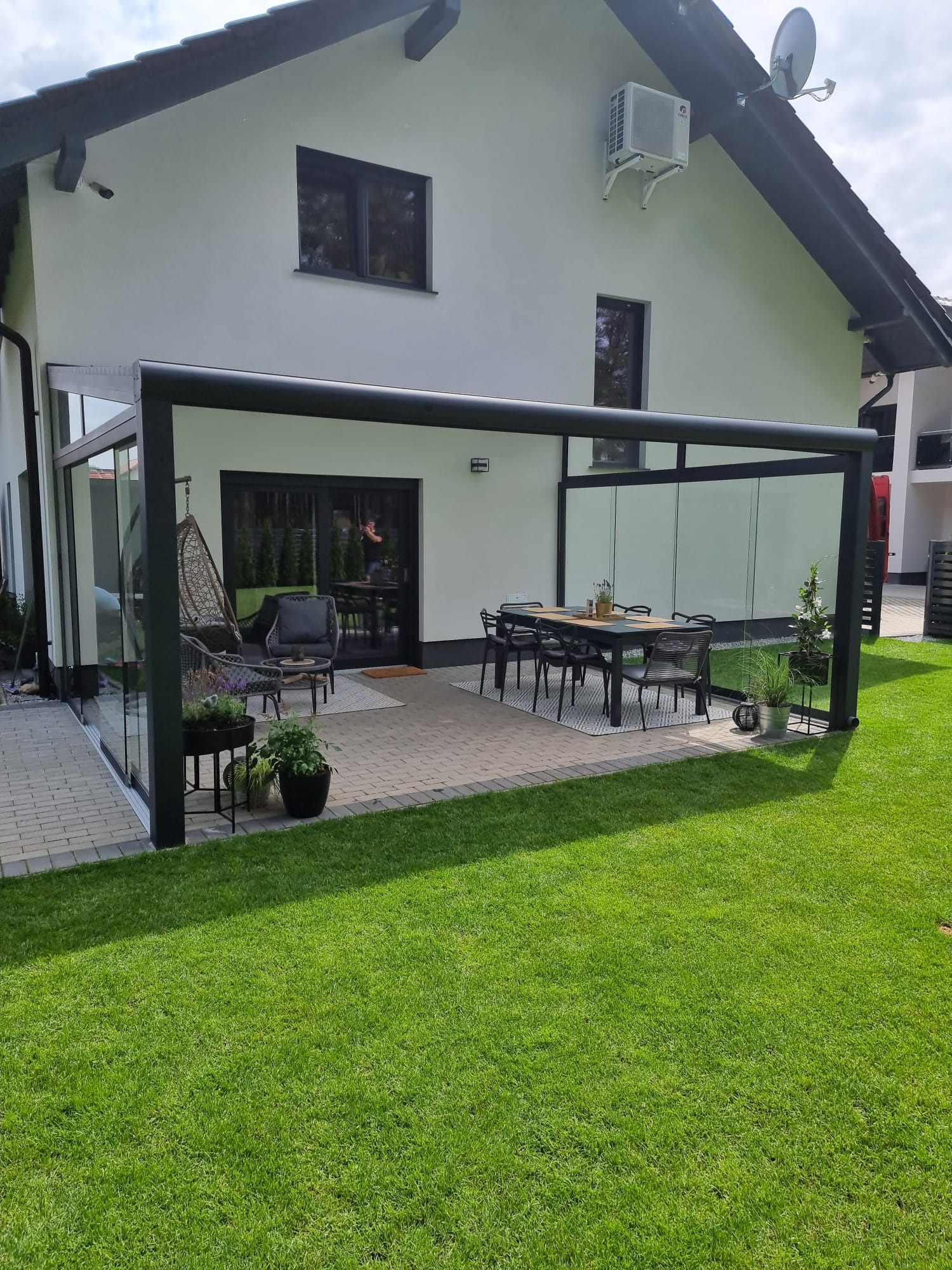 Nowoczesna pergola z przesuwnymi szklanymi panelami, przylegająca do białego domu z czarnymi oknami. Na tarasie stół z krzesłami, wiszący fotel i dekoracyjne rośliny w donicach.