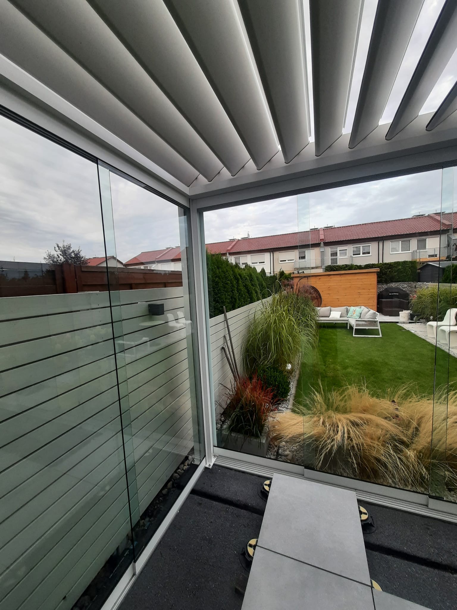 Nowoczesna, przeszklona pergola z regulowanymi lamelami dachowymi, widok na zadbany ogród z meblami wypoczynkowymi i roślinnością ozdobną.
