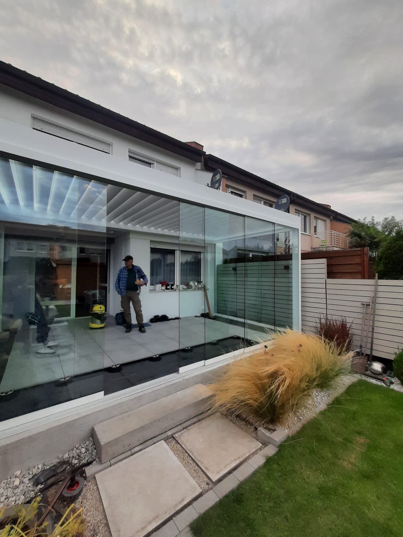 Zadaszony taras z przesuwnymi szklanymi ścianami, widoczny mężczyzna w trakcie prac wykończeniowych, na zewnątrz ozdobna trawa i fragment ogrodu.