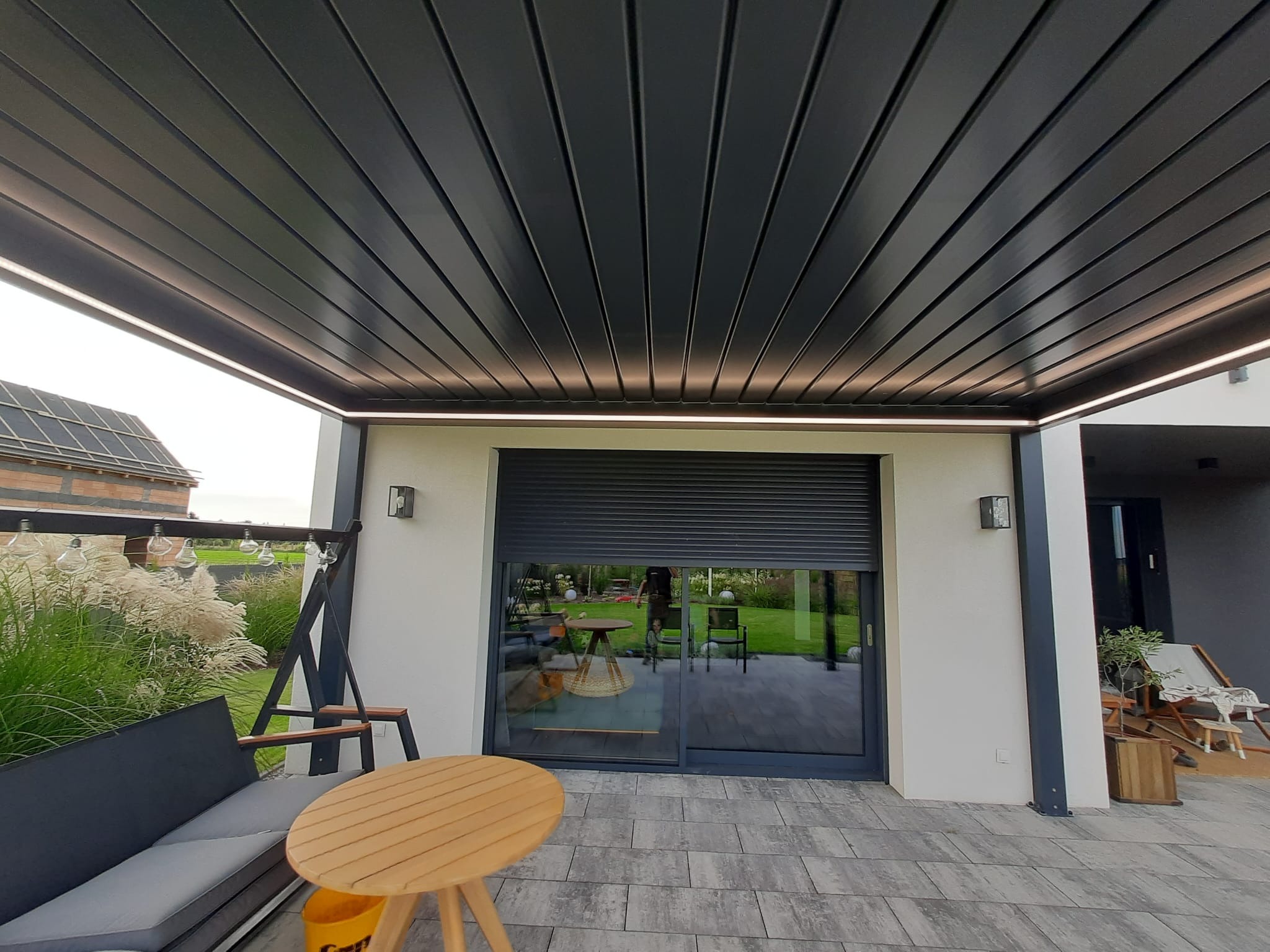Nowoczesna metalowa pergola z oświetleniem LED, widok z dołu na szare lamele, z przesuwnymi drzwiami i meblami ogrodowymi w tle.