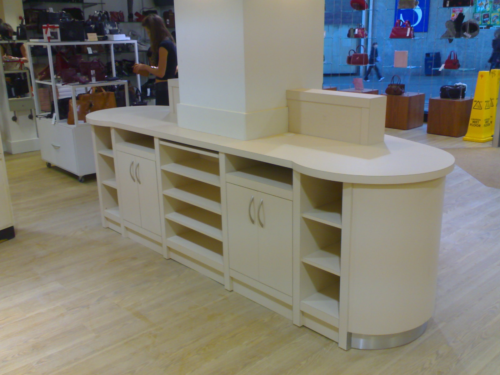 Jasny, zabudowany mebel sklepowy z półkami i szafkami, widoczny w tle sklep z torebkami i osoba przy półkach.