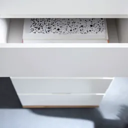 Biała komoda z wysuniętą szufladą, w której widać książki, widok z góry, delikatne oświetlenie.