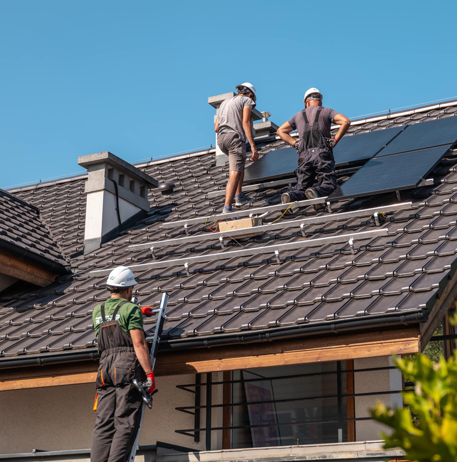 Montaż paneli na dachu: Trzech monterów w kaskach instaluje panele słoneczne na brązowym dachu z dachówki ceramicznej, jeden z nich stoi na drabinie z narzędziami, a dwaj pozostali pracują...