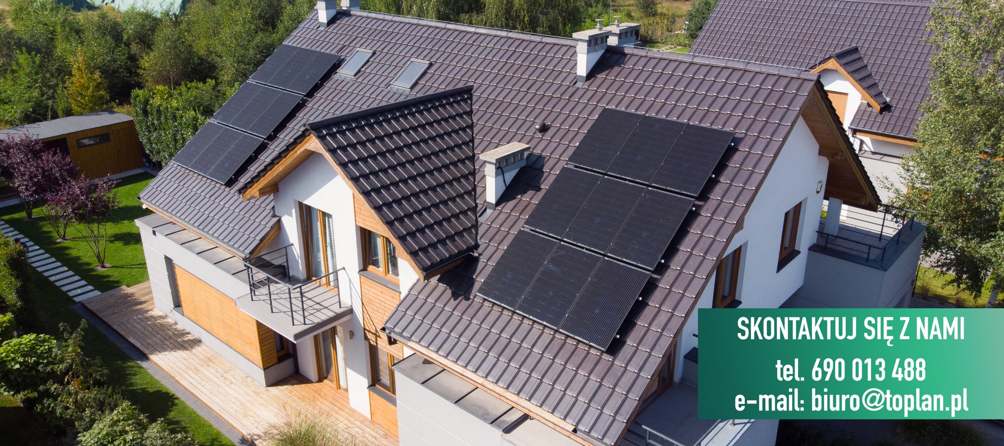 Widok z góry na dom z panelami słonecznymi na brązowym dachu, z widocznym balkonem i zielonym otoczeniem.