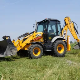 Żółto-czarna koparko-ładowarka JCB na zielonym terenie z widocznym stawem w tle, błękitne niebo.