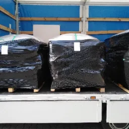 Ładunek trzech dużych, owiniętych czarną folią paczek na drewnianych paletach, umieszczony wewnątrz naczepy samochodu ciężarowego z niebieską plandeką.