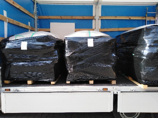 Ładunek trzech dużych, owiniętych czarną folią paczek na drewnianych paletach, umieszczony wewnątrz naczepy samochodu ciężarowego z niebieską plandeką.