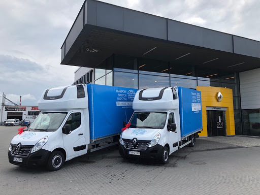 Dwa białe busy dostawcze Renault z niebieską plandeką, zaparkowane przed salonem samochodowym Renault, z dekoracyjnymi czerwonymi kokardami na lusterkach.