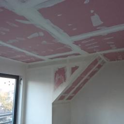 FHU Dawid Bigos - Montaż płyt karton-gips na suficie poddasza z widocznymi profilami i szpachlowaniem łączeń, drewniana belka oparta o ścianę, okno balkonowe w tle.