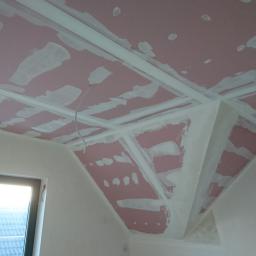 FHU Dawid Bigos - Poddasze z ukośnym sufitem z płyt kartonowo-gipsowych, wstępnie szpachlowanych na różowo, widoczne profile montażowe i fragment okna dachowego z widokiem na dach sąsiedniego budynku.