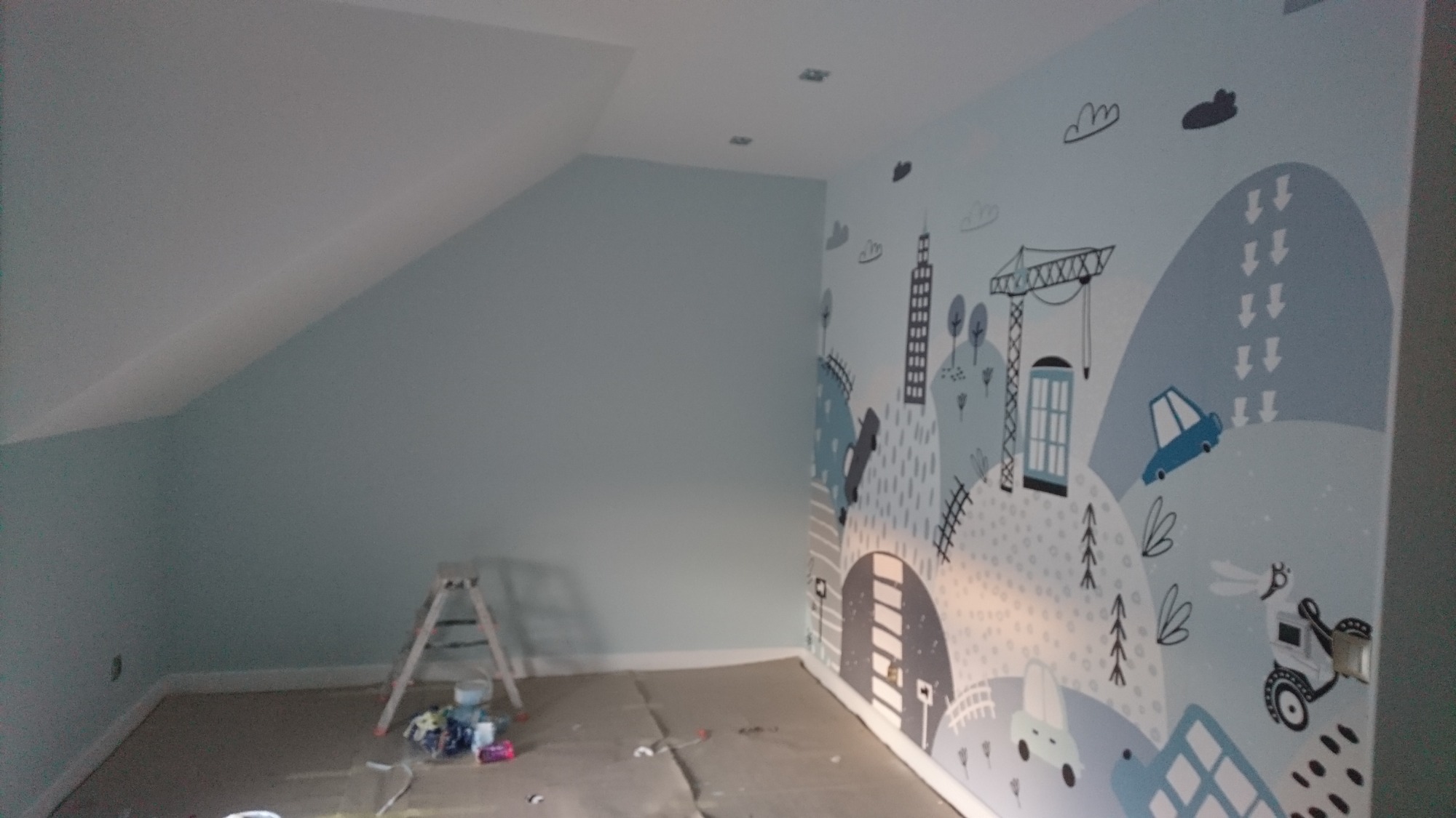 Pokój w Mikorzynie po malowaniu z motywem miejskim na ścianie, widoczna drabina i akcesoria malarskie, przygotowany do użytkowania.