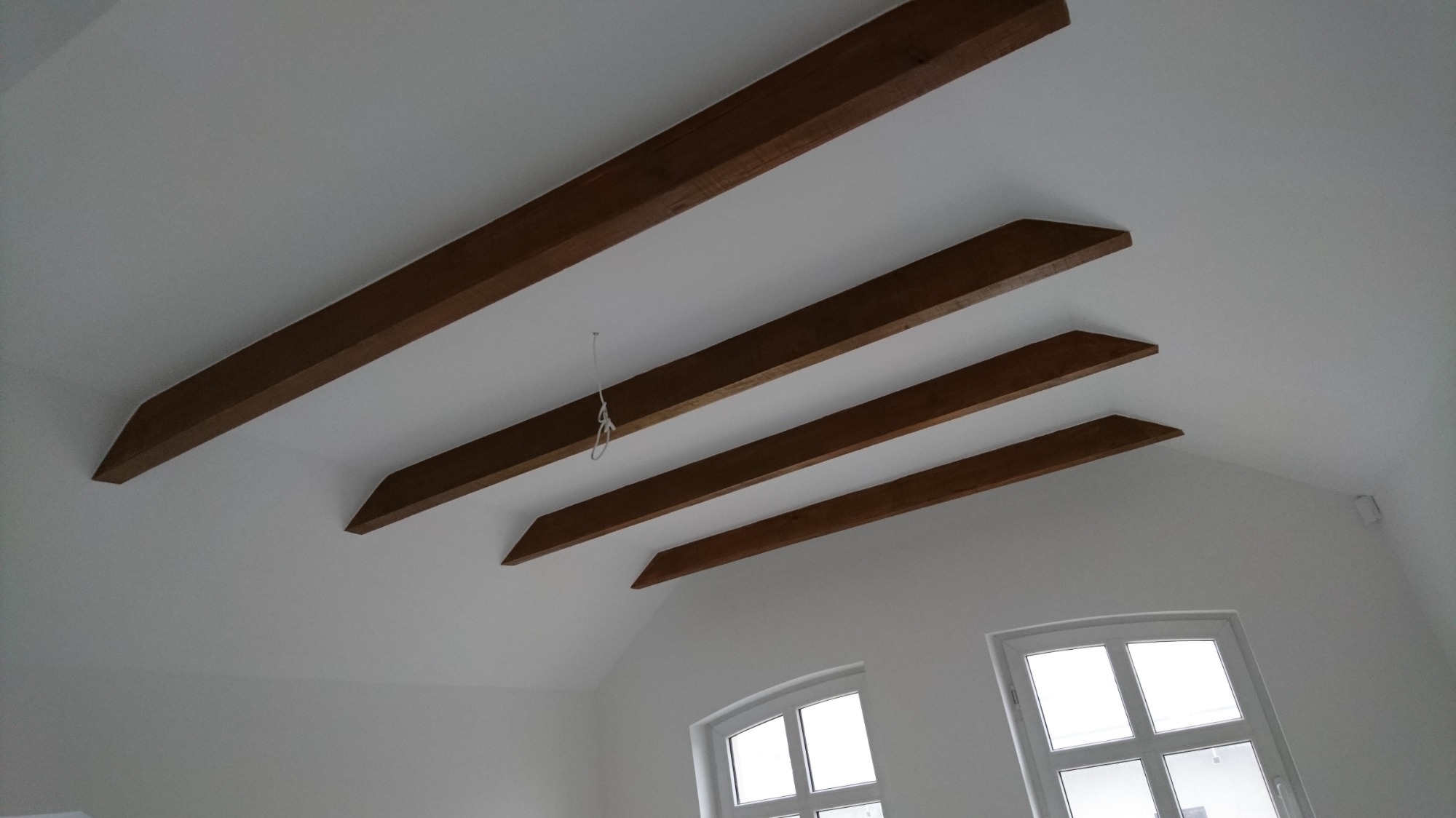 Białe wnętrze poddasza z czterema równoległymi, brązowymi belkami stropowymi o trapezoidalnych zakończeniach, widoczne okno dachowe z białymi ramami i zwisający sznurek.