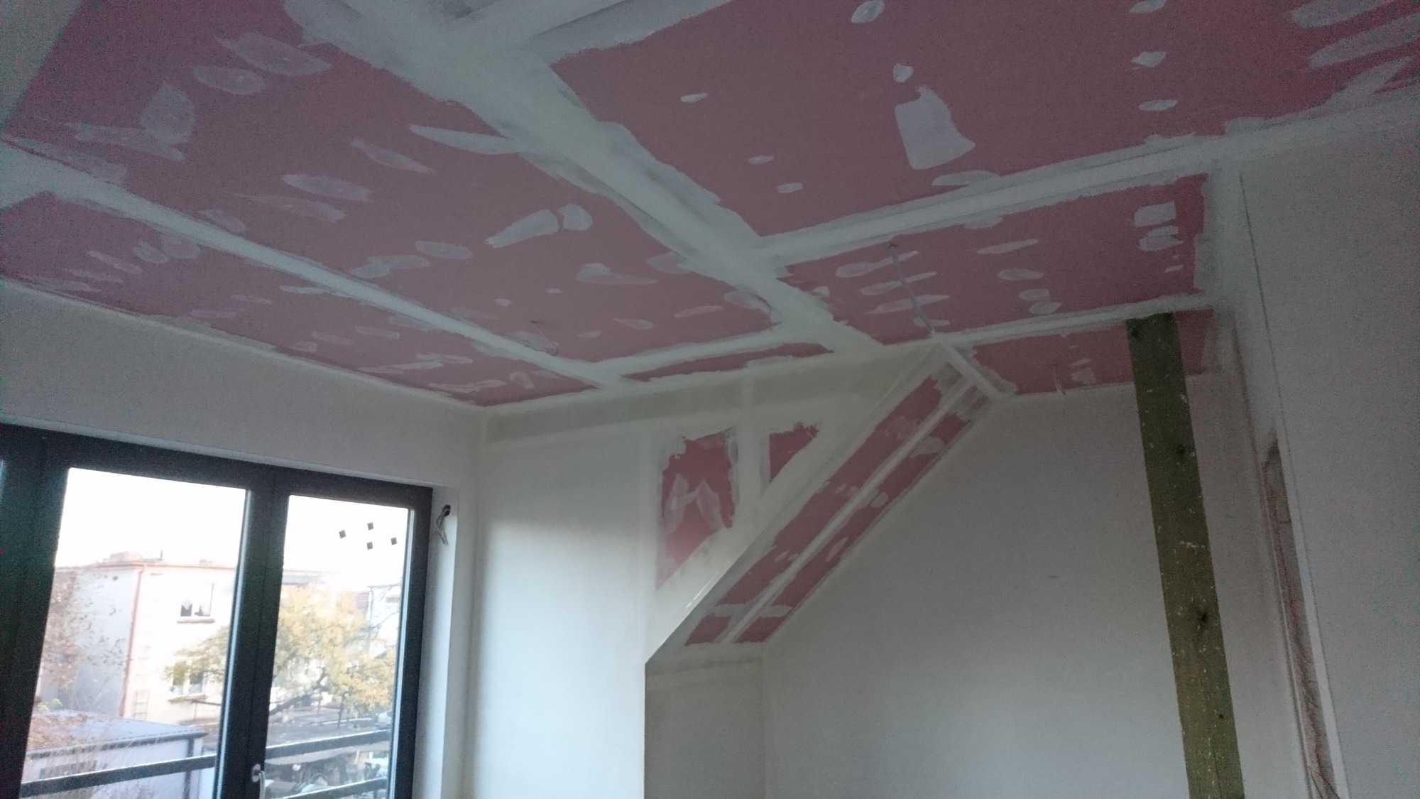 Montaż płyt karton-gips na suficie poddasza z widocznymi profilami i szpachlowaniem łączeń, drewniana belka oparta o ścianę, okno balkonowe w tle.