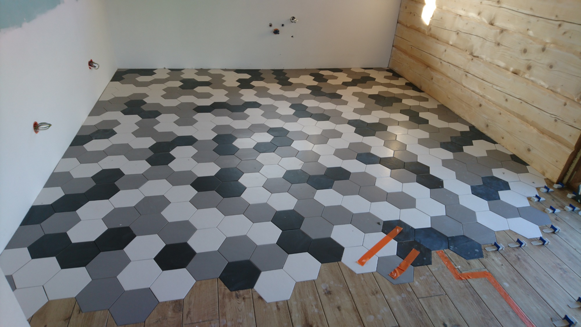 Układanie podłogi z heksagonalnych płytek w odcieniach szarości i bieli, widoczne elementy dystansowe i taśma malarska na drewnianej podłodze, surowa ściana z bali w tle.