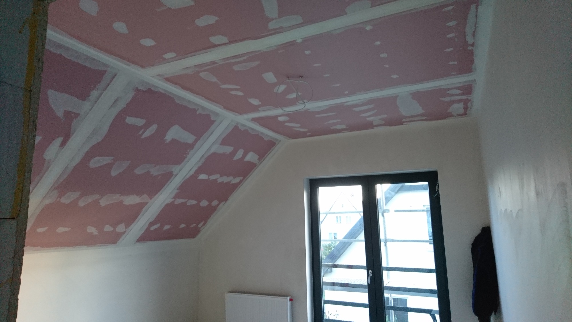 Poddasze z zamontowaną płytą kartonowo-gipsową na skośnym suficie, widoczne łączenia płyt wypełnione masą szpachlową, okno dachowe w tle.