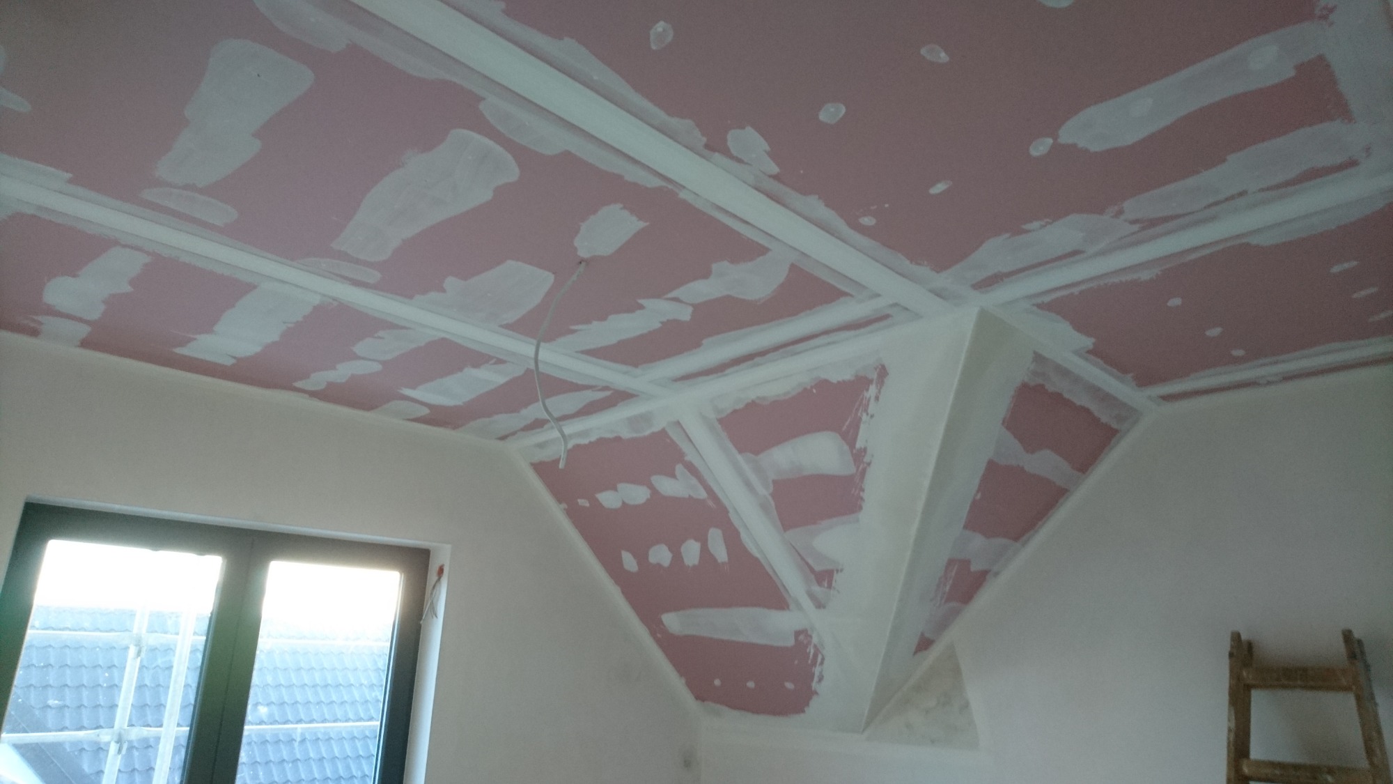 Poddasze z ukośnym sufitem z płyt kartonowo-gipsowych, wstępnie szpachlowanych na różowo, widoczne profile montażowe i fragment okna dachowego z widokiem na dach sąsiedniego budynku.