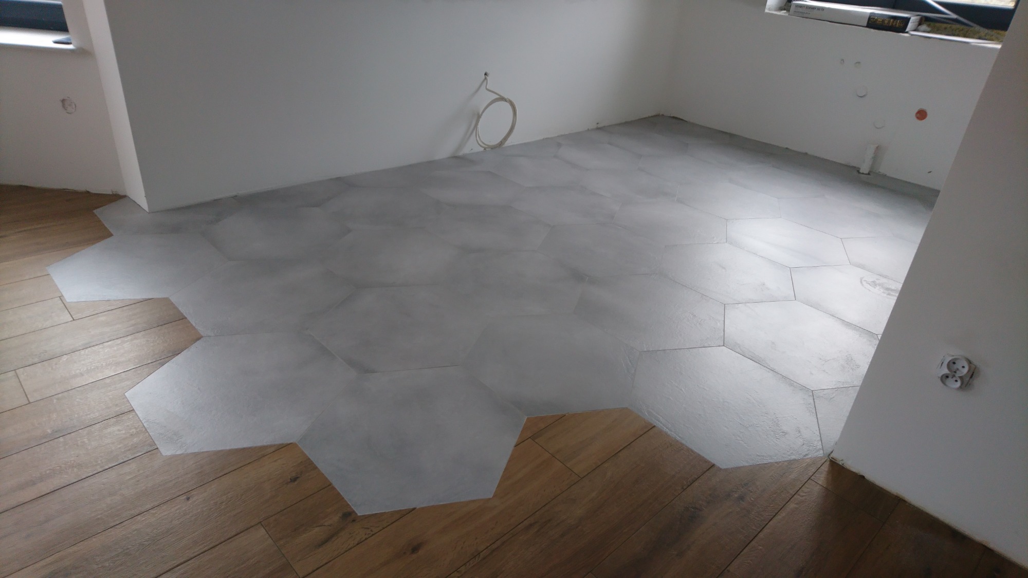 Pokój w trakcie wykańczania z szarymi, heksagonalnymi płytkami częściowo ułożonymi na podłodze, obok widoczna drewniana podłoga.
