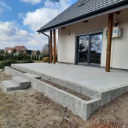 Wykończony taras z szarej kostki na dwóch poziomach z niskimi murkami, schody z kostki, dom z szarym dachem w tle, błękitne niebo z chmurami.