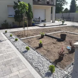 Przygotowany teren pod ogród z ułożoną szarą kostką brukową, obrzeża rabat wysypane białym grysem, sadzonki w doniczkach czekają na posadzenie.