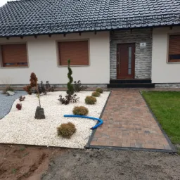 Ogród przed domem z ścieżką z kostki, rabatą z białego kamienia, ozdobnymi krzewami i niebieskim wężem drenażowym.