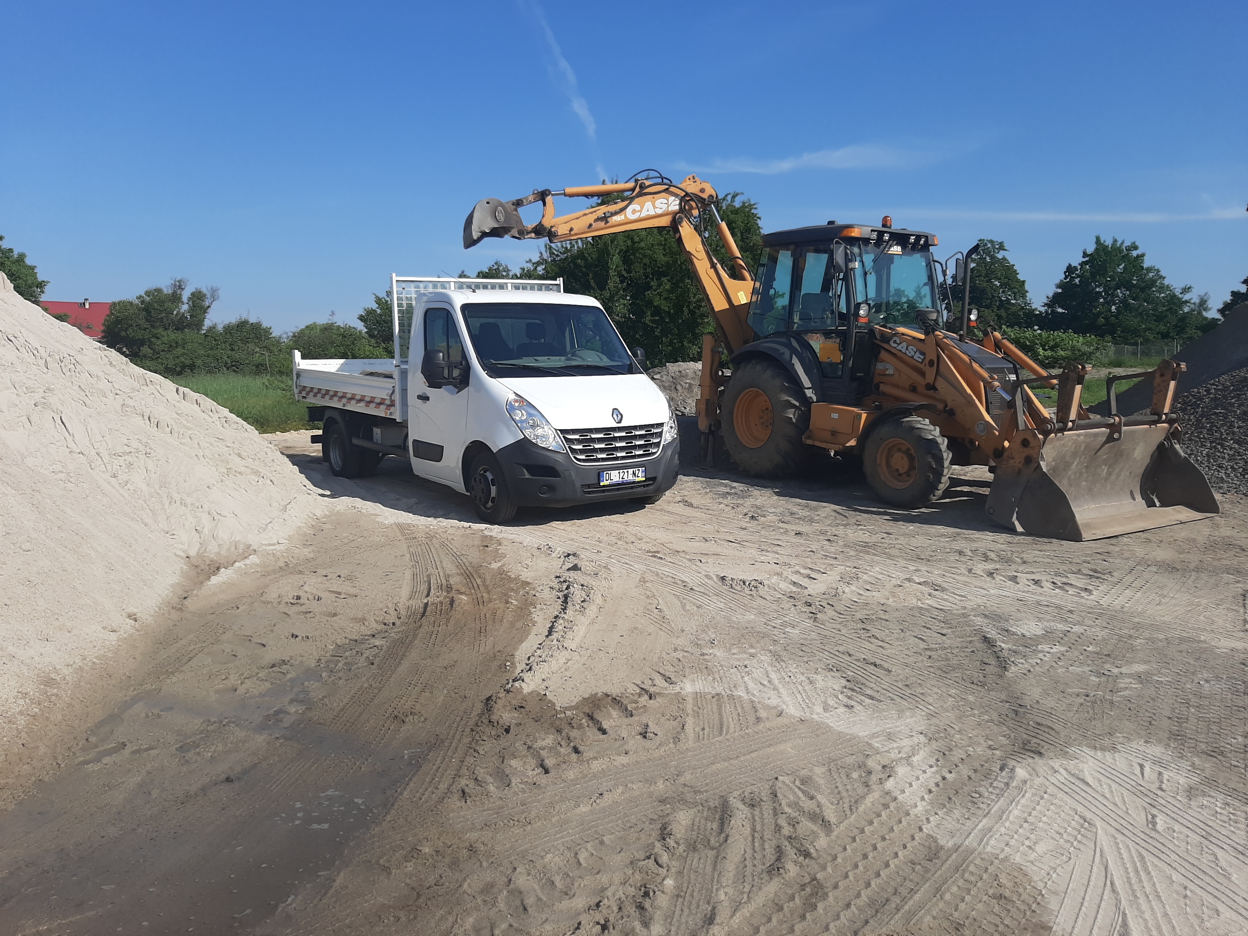 Biała wywrotka Renault i koparko-ładowarka Case na placu budowy z hałdą piasku w tle, błękitne niebo.