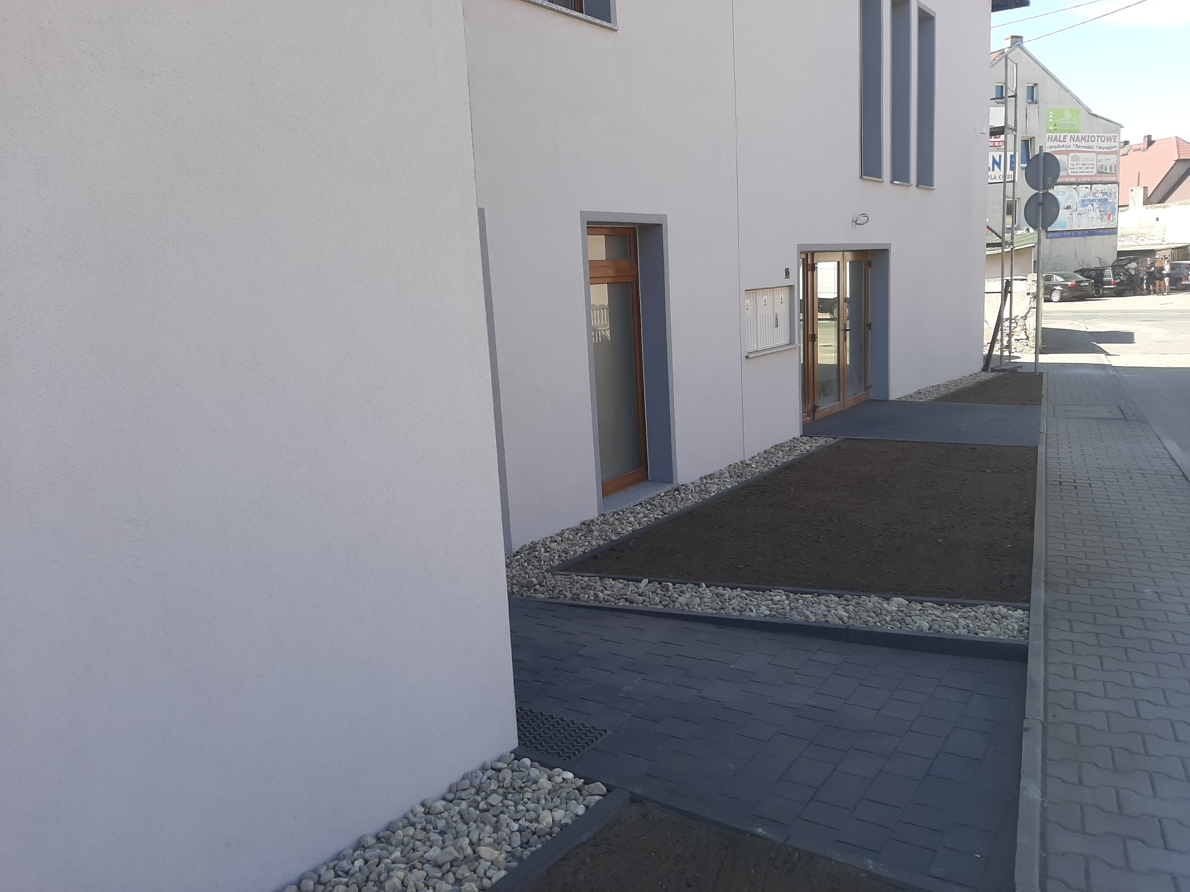 Nowoczesna aranżacja terenu przed budynkiem: ciemna kostka brukowa, rabaty z ziemią obłożone białym żwirem, jasna elewacja budynku.