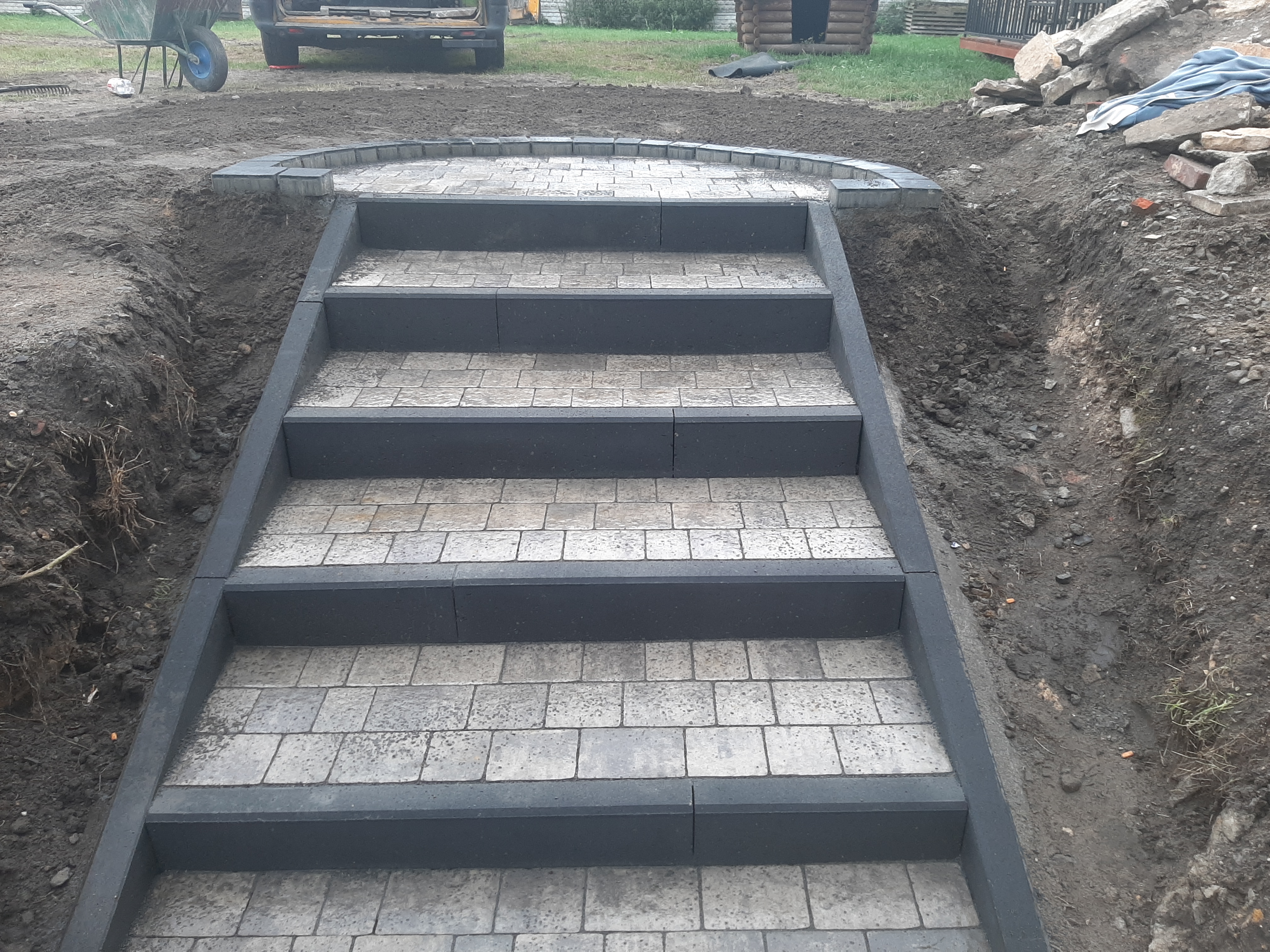 Schody zewnętrzne z szarej kostki brukowej, obramowane ciemnoszarym obrzeżem, prowadzące w górę, z widocznymi śladami prac ziemnych po bokach.