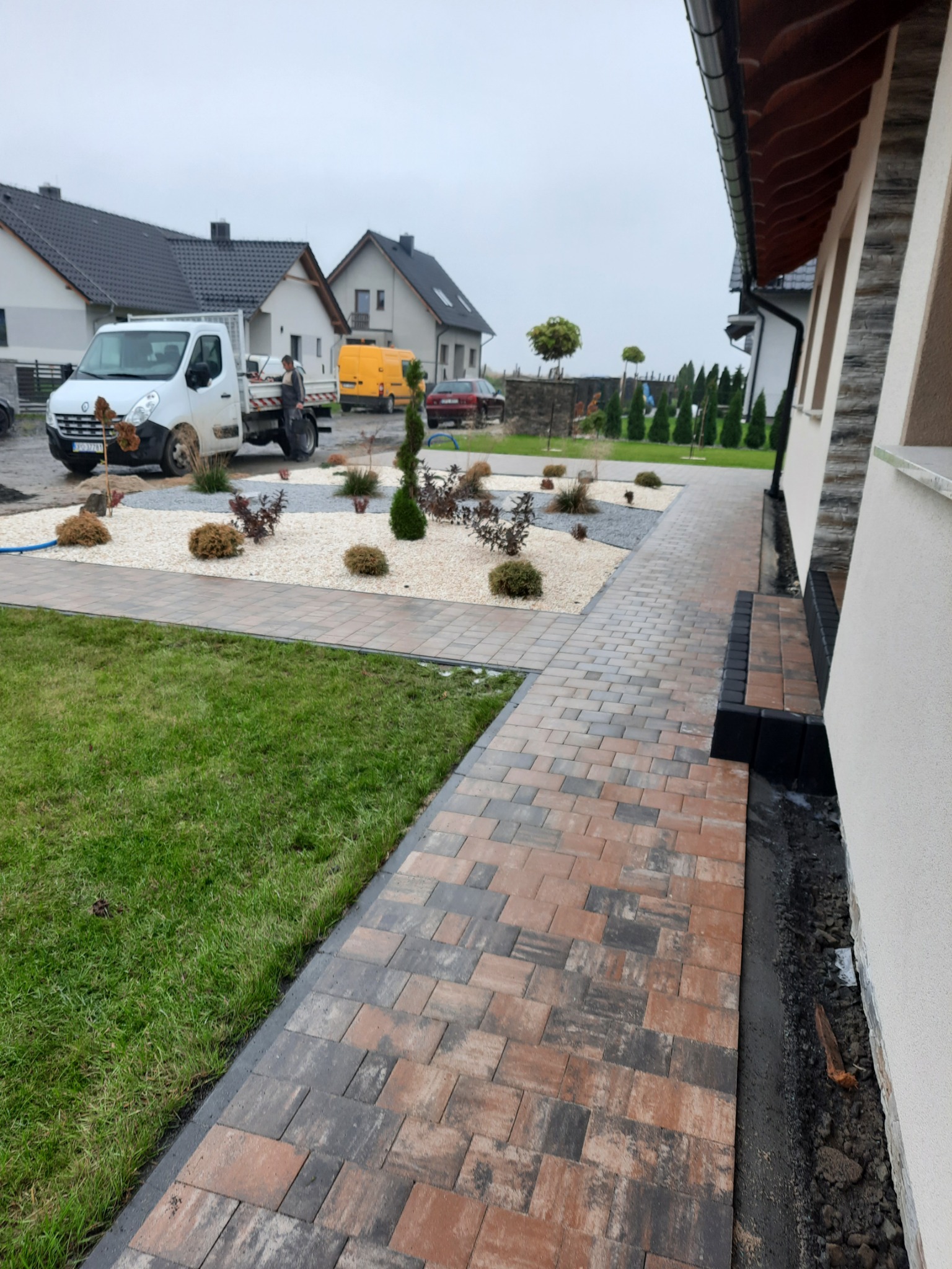 Ułożona kostka brukowa w odcieniach brązu i szarości tworząca ścieżkę obok trawnika i rabaty z białym żwirem oraz niskimi krzewami, w tle zaparkowany biały samochód dostawczy i inne domy.