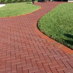 Czerwona ścieżka z cegły ułożona w jodełkę, obrzeża z pojedynczych cegieł, trawnik po bokach, w oddali ławka i donica.