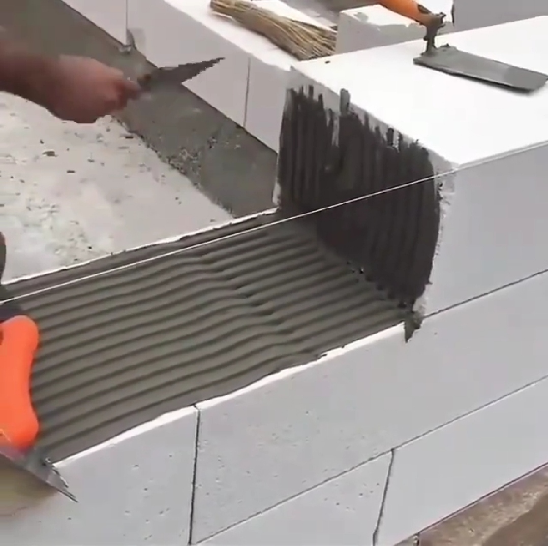 Wznoszenie ściany z białych bloczków, z nałożoną warstwą zaprawy i napiętym sznurkiem murarskim, widoczna dłoń murarza z kielnią.