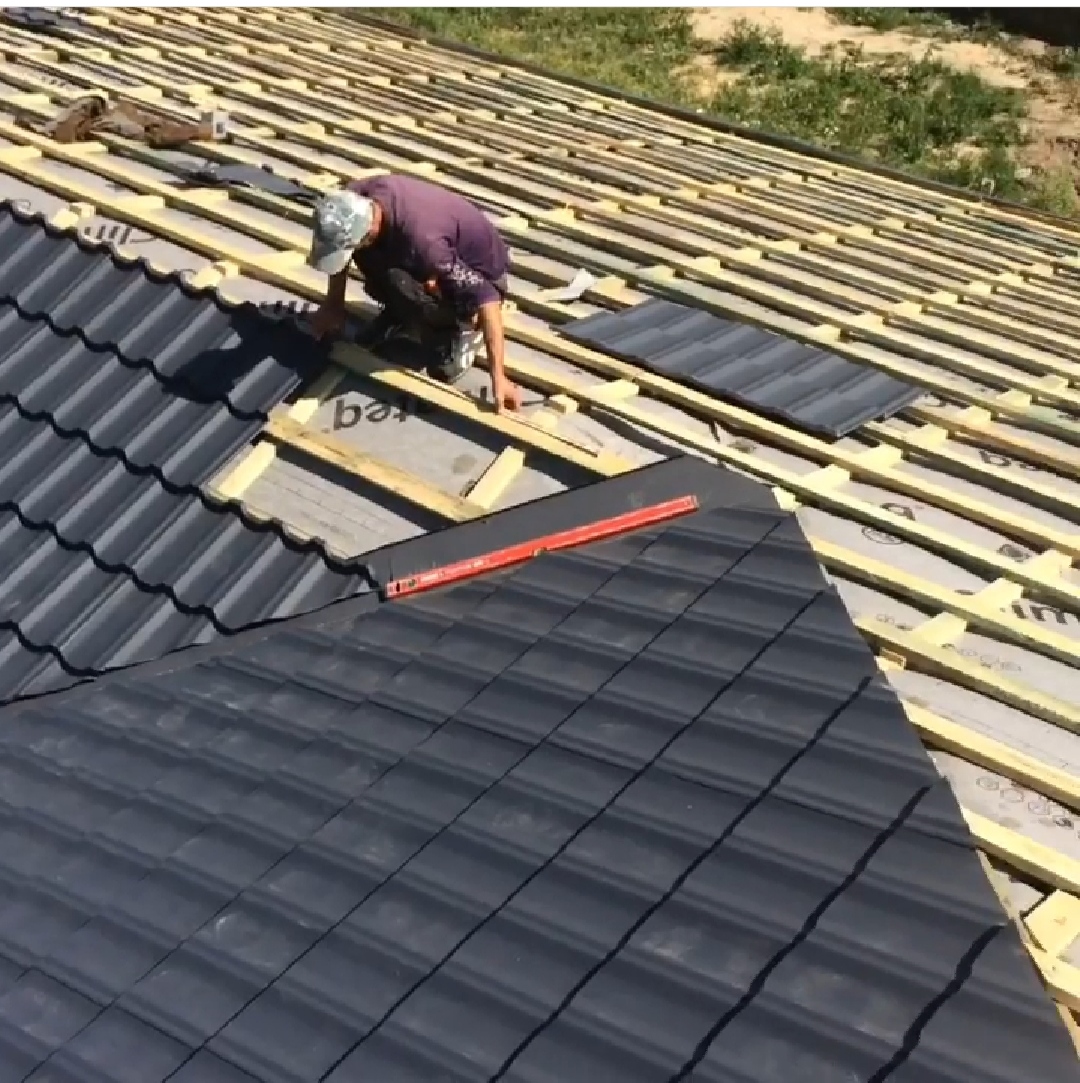 Pracownik w czapce układa ciemne dachówki na drewnianej konstrukcji dachu, widoczny poziomica w kolorze pomarańczowym.