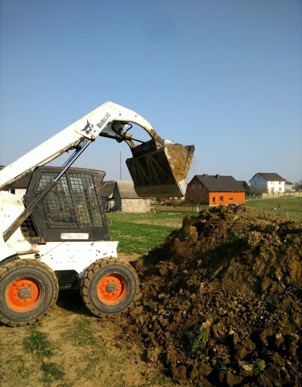 Biała miniładowarka Bobcat z pomarańczowymi felgami nabiera ziemię z pryzmy, w tle zabudowa jednorodzinna i błękitne niebo.