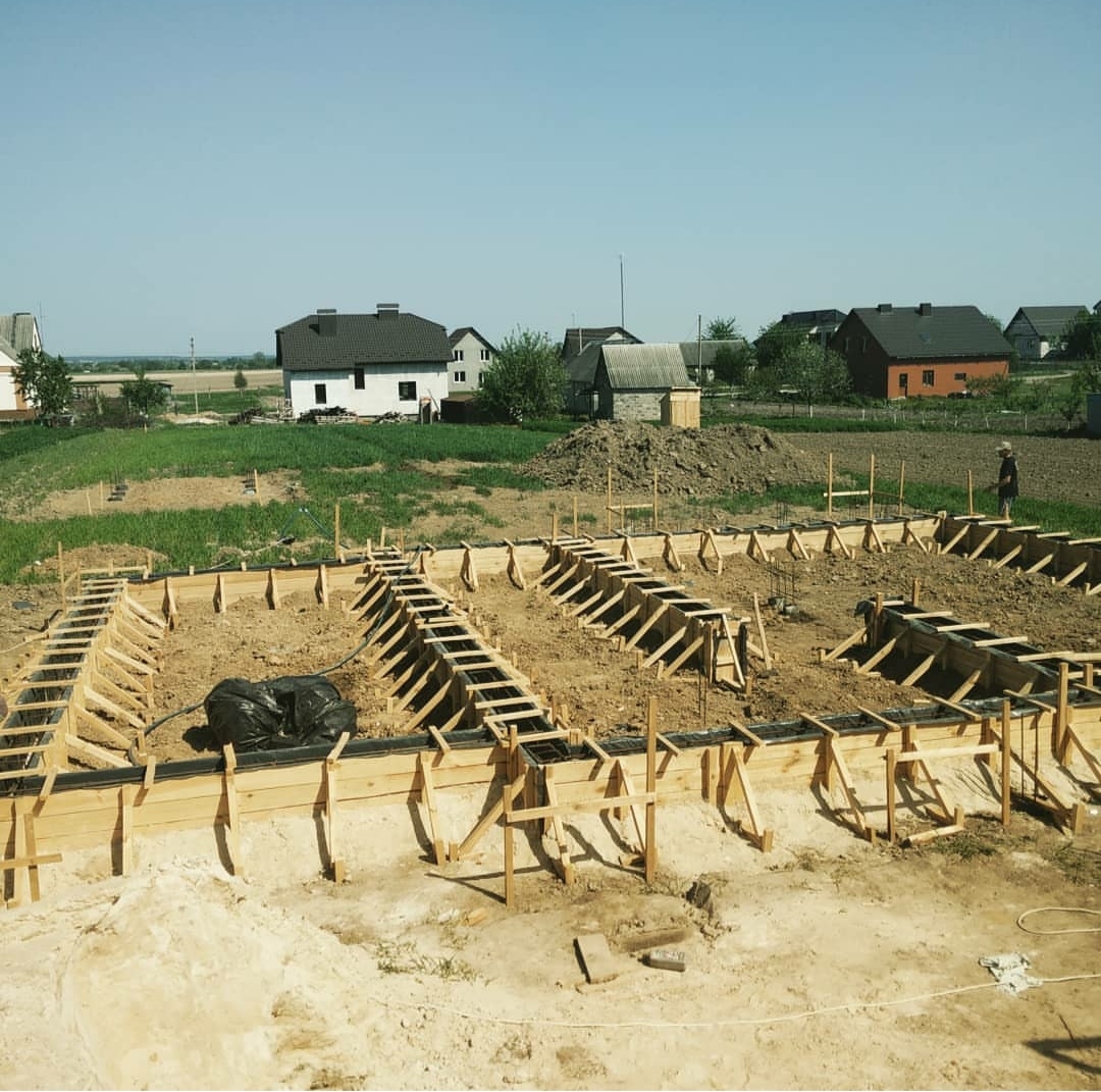 Szeroki kadr na drewniane szalunki fundamentów domu jednorodzinnego, z widocznymi elementami zbrojenia i czarną folią izolacyjną, w tle zabudowa wiejska i błękitne niebo.