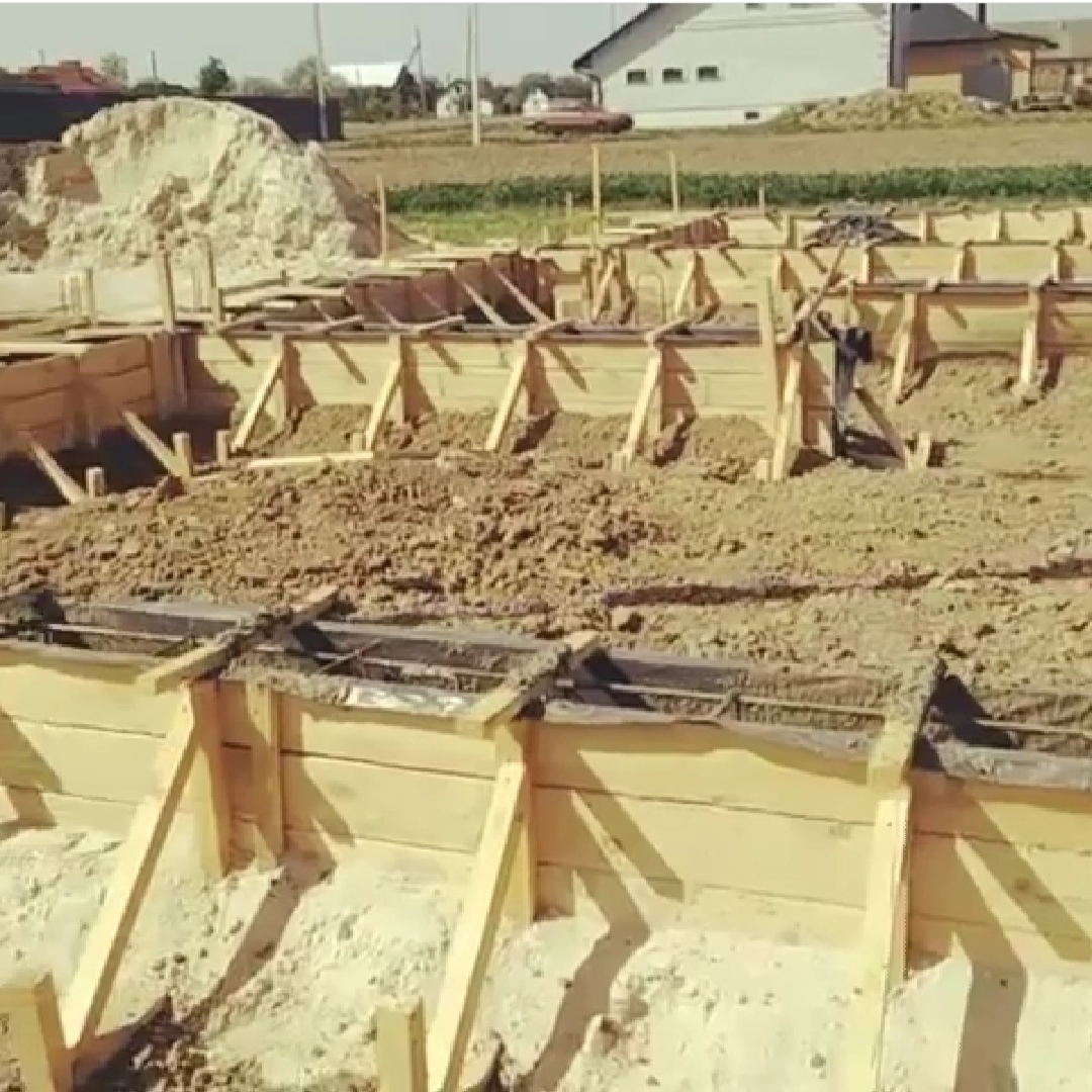 Drewniany szalunek fundamentów domu jednorodzinnego, wypełniony ziemią, z widocznym zbrojeniem, w tle hałda ziemi i budynek.