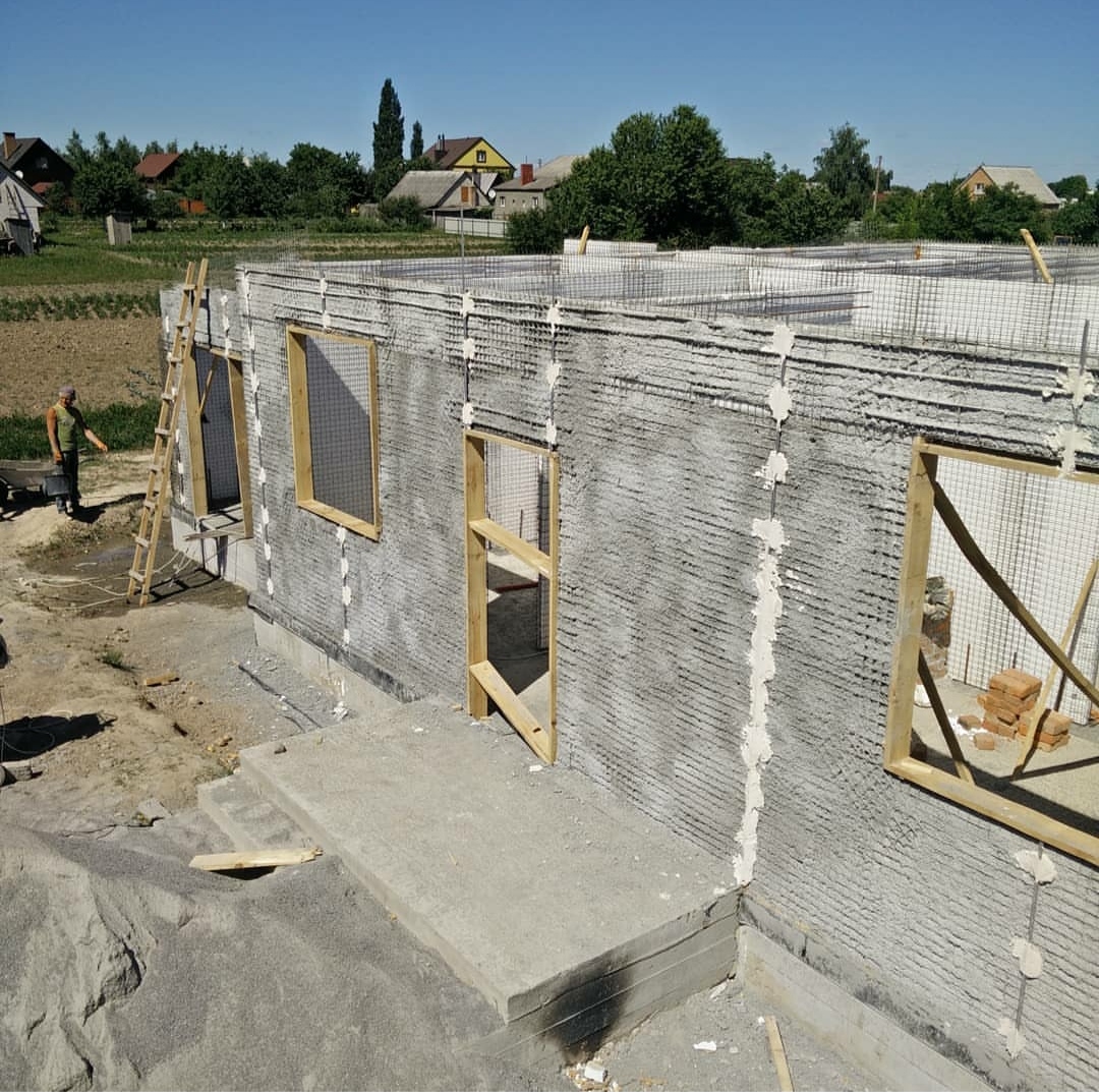 Wznoszenie ścian parterowego domu z szarego betonu komórkowego z widocznymi otworami okiennymi i drzwiowymi, drewnianymi ramami, siatką zbrojeniową i pracownikiem w oddali.