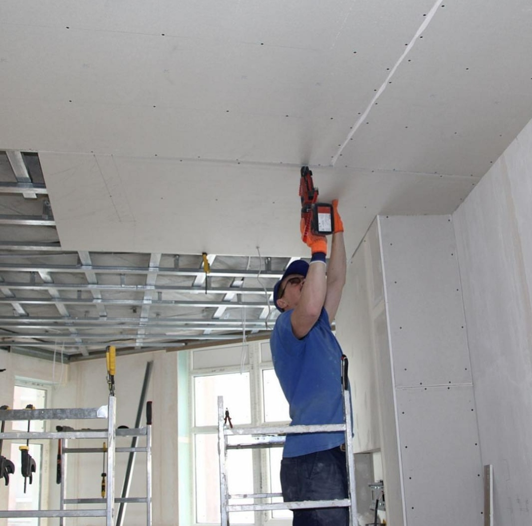 Pracownik w niebieskiej koszulce i pomarańczowych rękawicach montuje płytę karton gips na suficie za pomocą specjalistycznego narzędzia, widoczna metalowa konstrukcja sufitu podwieszanego i drabina.