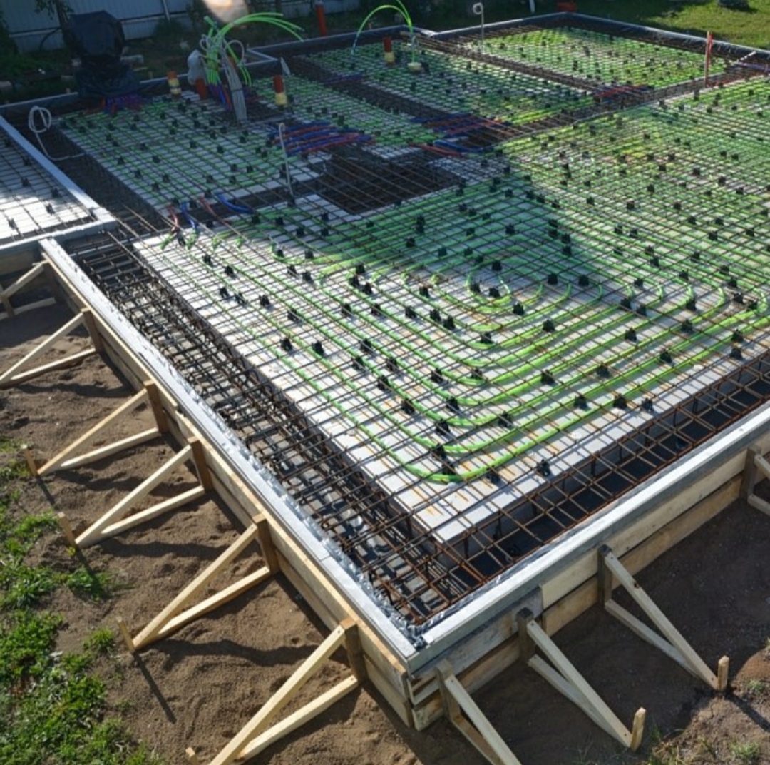 Przygotowanie płyty fundamentowej z ogrzewaniem podłogowym: zbrojenie stalowe i rury grzewcze ułożone w drewnianym szalunku, widoczne wsporniki i częściowo wylany beton.